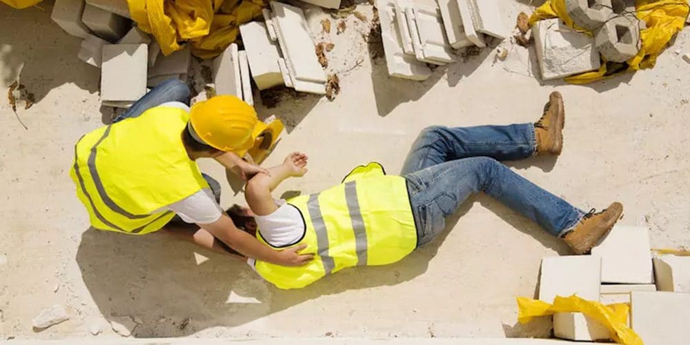 A workman in a yellow vest at a construction site is laying on the ground after being injured. A fellow workman is trying to help.