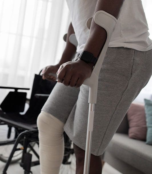 A man with a medically wrapped right knee and lower leg struggles to walk with arm crutches.