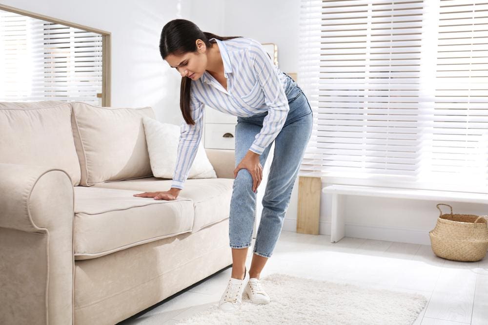 A woman is struggling to get up from the couch and walk because of knee pain.