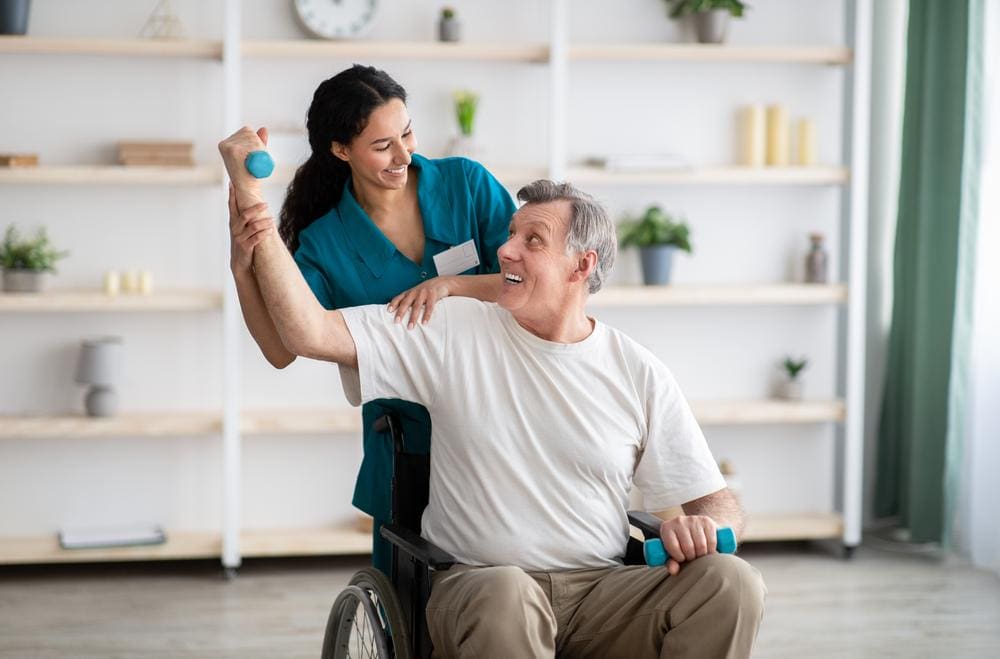 An elderly man is doing arm exercises with the assistance of a physical therapist. 
