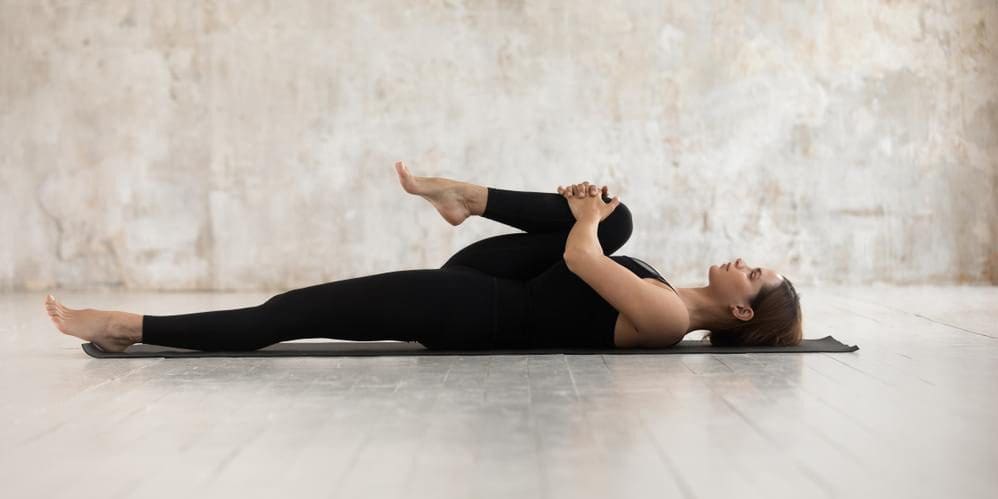 A woman is lying on her back and stretching by pulling her knee towards her chest.