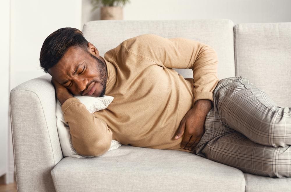 A man is laying on the couch holding his stomach because of abdominal pain. 