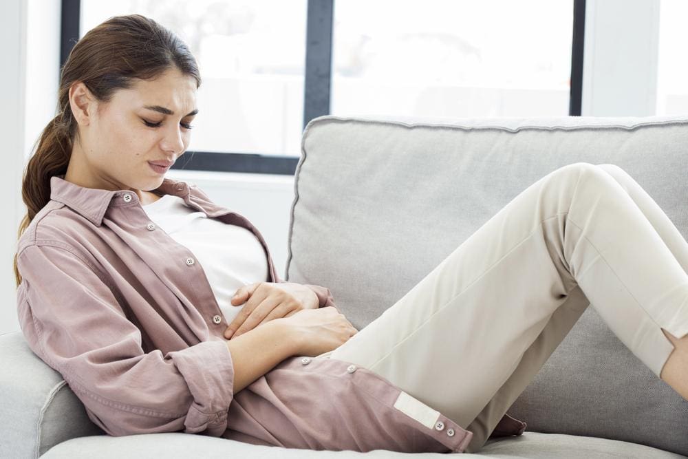 A woman is on the couch clutching her stomach because she is suffering from abdominal pain.