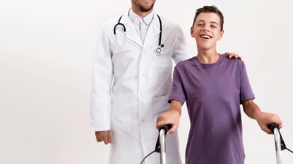 Teenage boy with cerebral palsy standing with walker with doctor behind with hand on his shoulder.