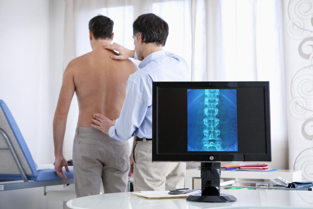 A doctor is examining the spine of male patient in a doctor's office with X-ray image on computer screen.