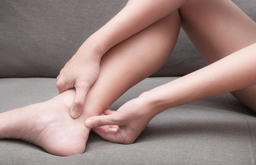 A woman is sitting on the couch and rubbing her ankle because she has achilles tendonitis. 