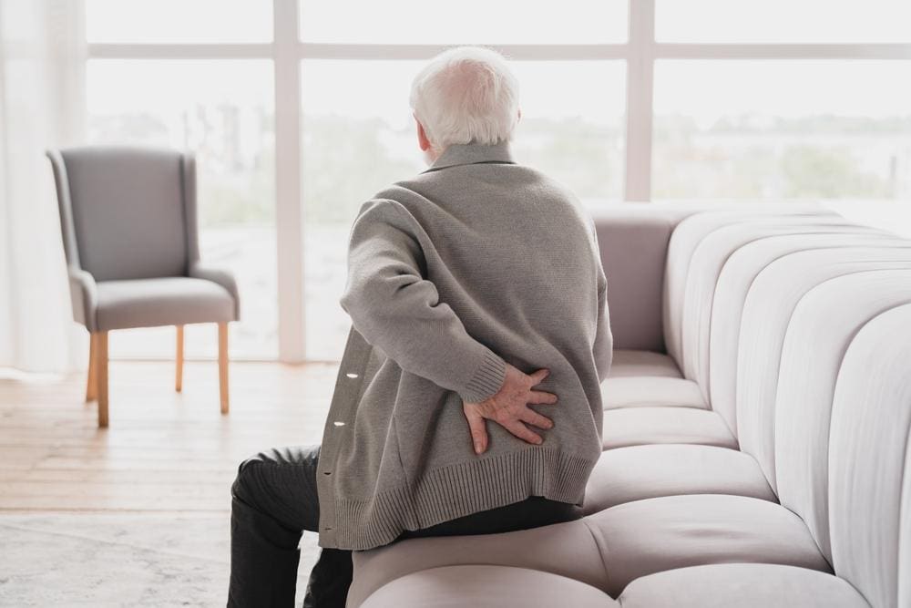 An elderly man is sitting on the couch, and not getting up because of back pain.
