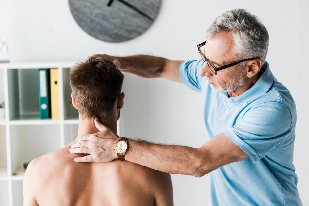 Chiropractor with glasses adjusting neck of young male patient.
