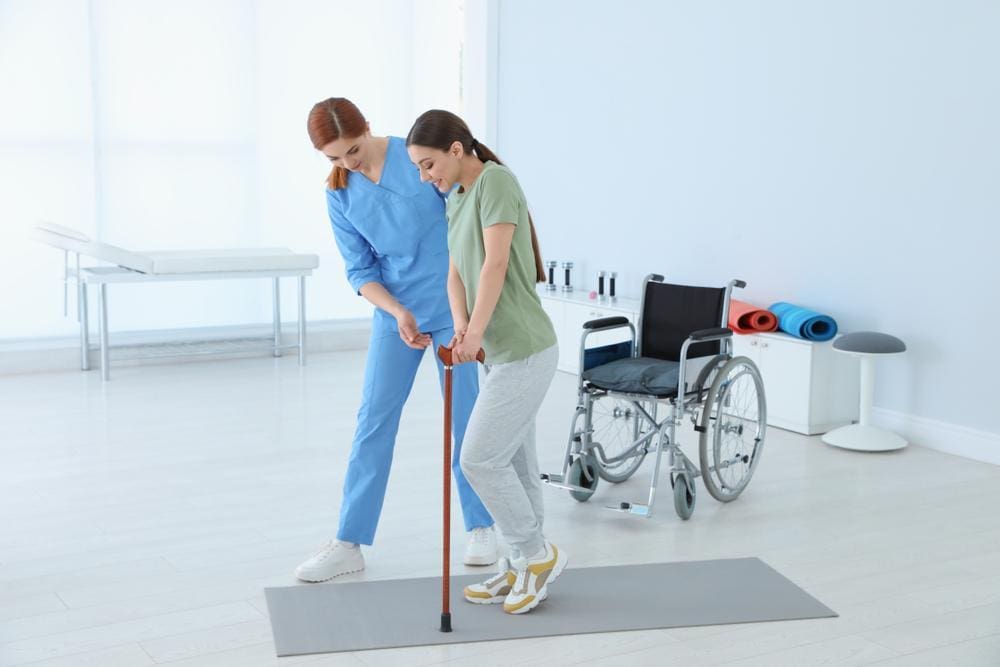 Physiotherapist helping young female patient walk with a cane.

