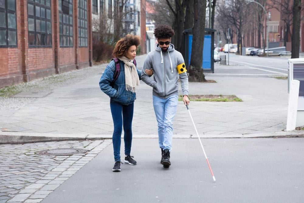 Young woman helping visually impaired man with white cane cross the street.
