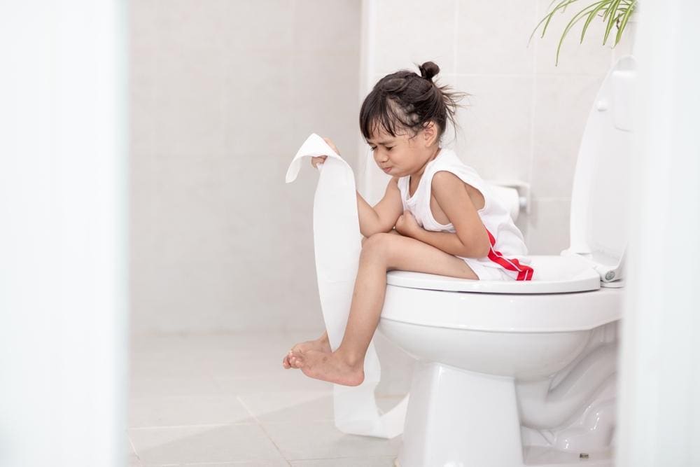 Child sitting on toilet holding length of toilet paper struggling with constipation.