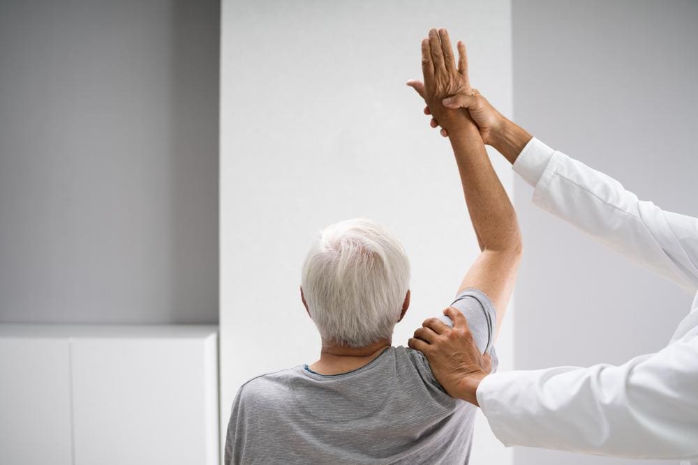 Senior patient with arm held into the air by chiropractor during treatment for frozen shoulder. 
