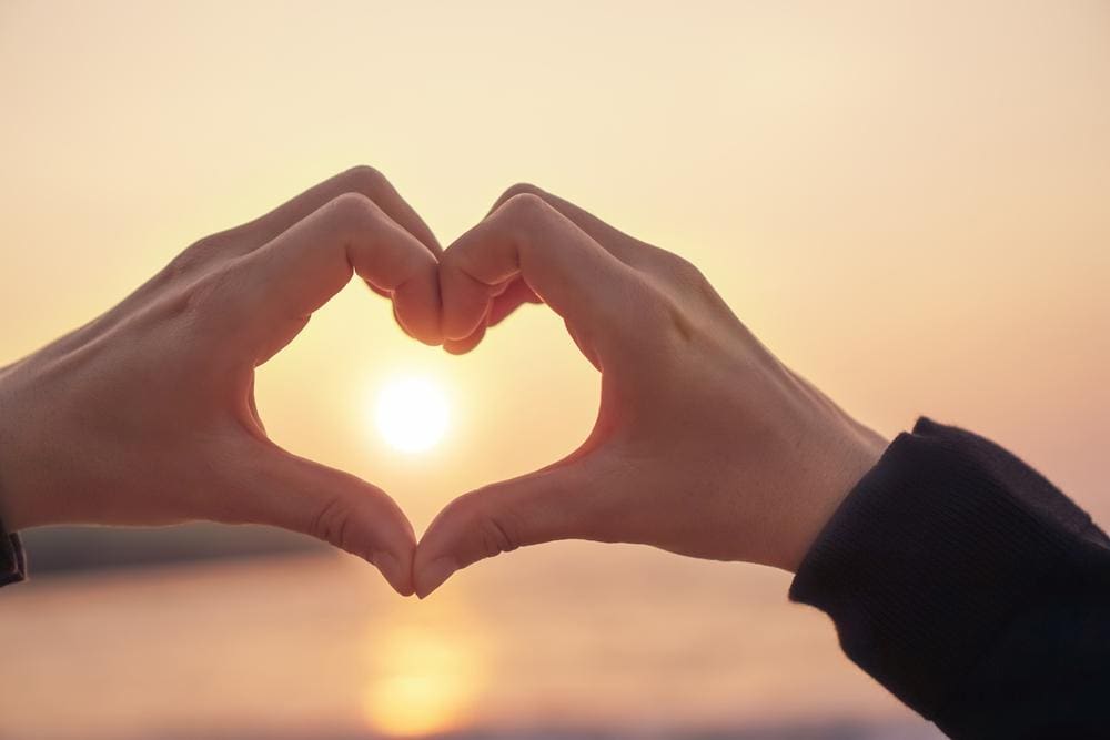 Woman's hands held in shape of heart surrounding the setting sun.
