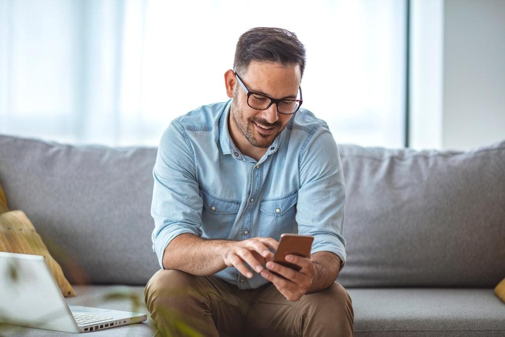 Man with glasses sitting on sofa, hunched over using a smartphone.
