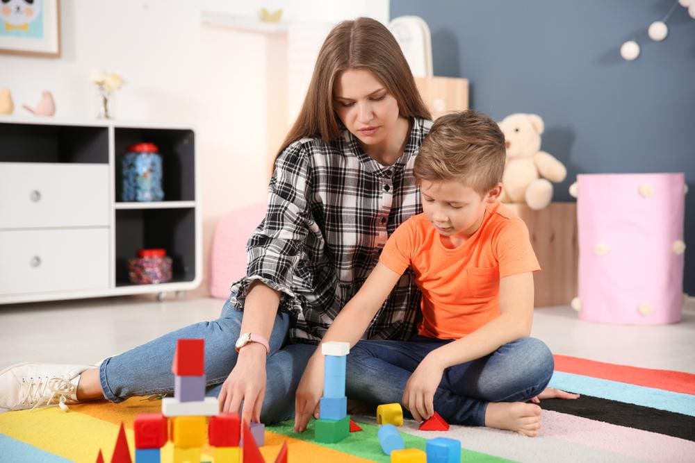 A young mother sitting with her autistic son, playing with blocks on mat.
