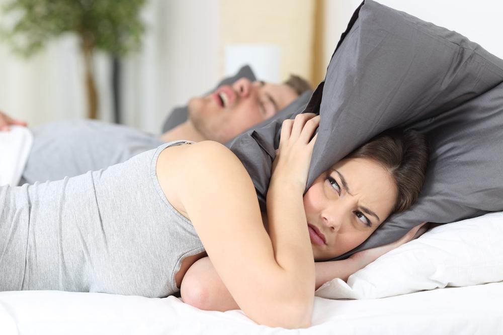 Woman holding a pillow over her ears while man sleeping beside her snores.
