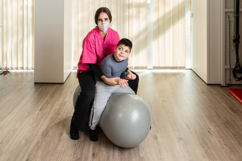 Physiotherapist in scrubs and mask sitting on exercise ball with boy with cerebral palsy.
