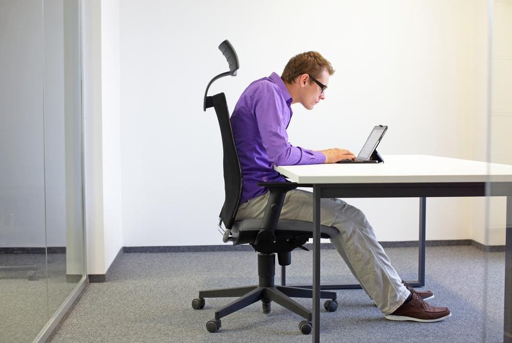 Man sitting hunched over at desk in office chair typing on computer.


