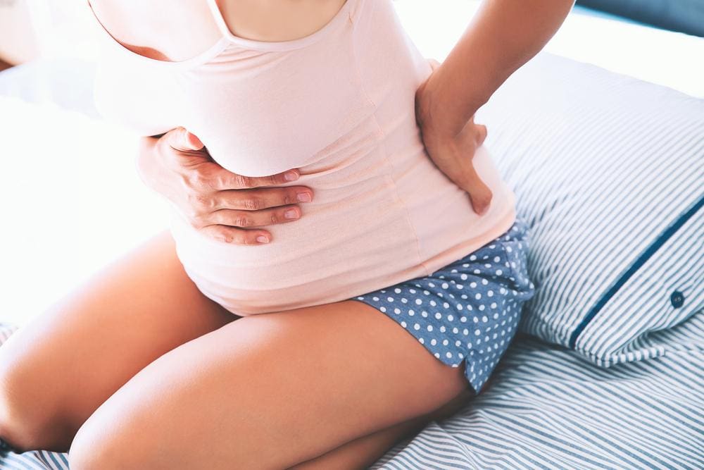 Pregnant woman kneeling on bed holding her lower back and stomach.
