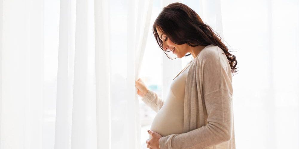 Pregnant woman smiling and holding her belly while standing in front of window.