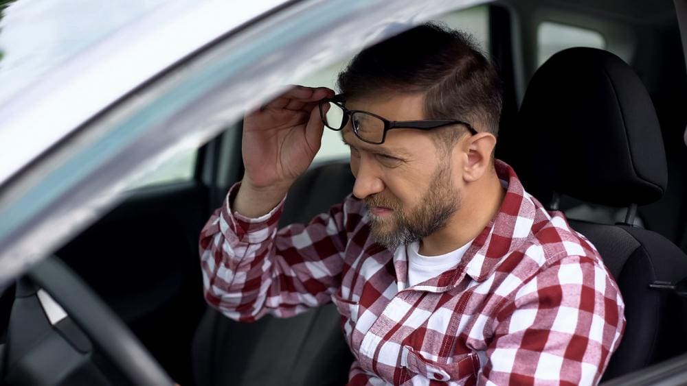 Man sitting in driver's seat of car squinting while lifting up glasses.