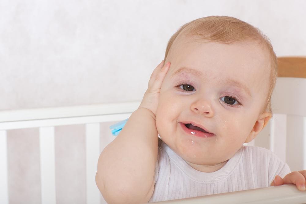 Baby girl standing in in crib holding her ear.
