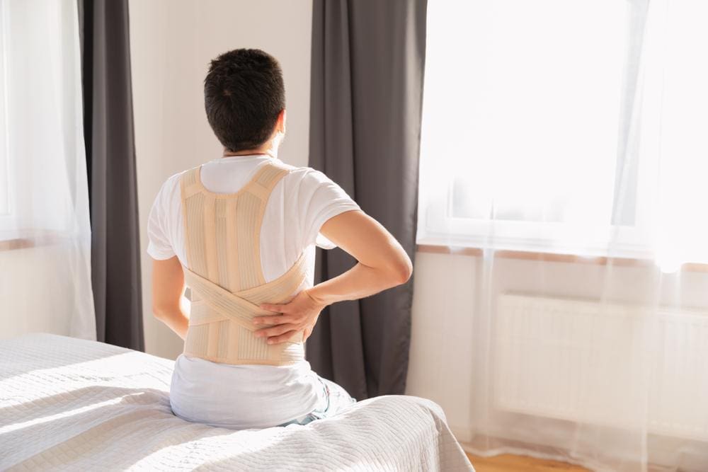 Young man sitting on the edge of a bed wearing a back brace.