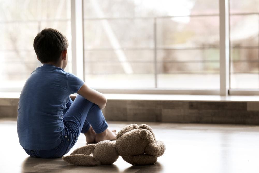 Young boy sitting on the floor next to a teddy bear, looking out the window.