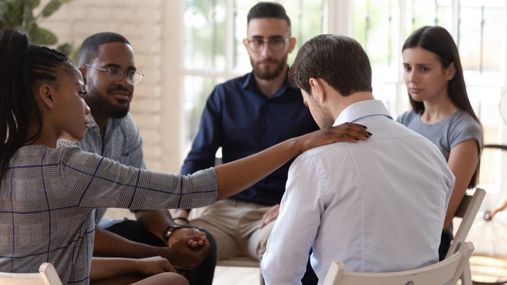 A man attending a support group for addiction recovery is comforted by his peers.
