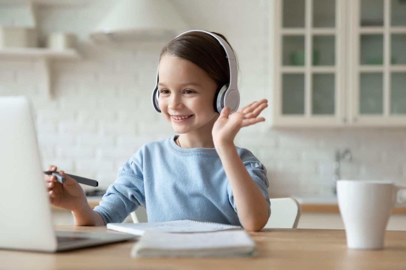 School-aged girl wearing headphones and waving at a laptop, attending an online therapy session.