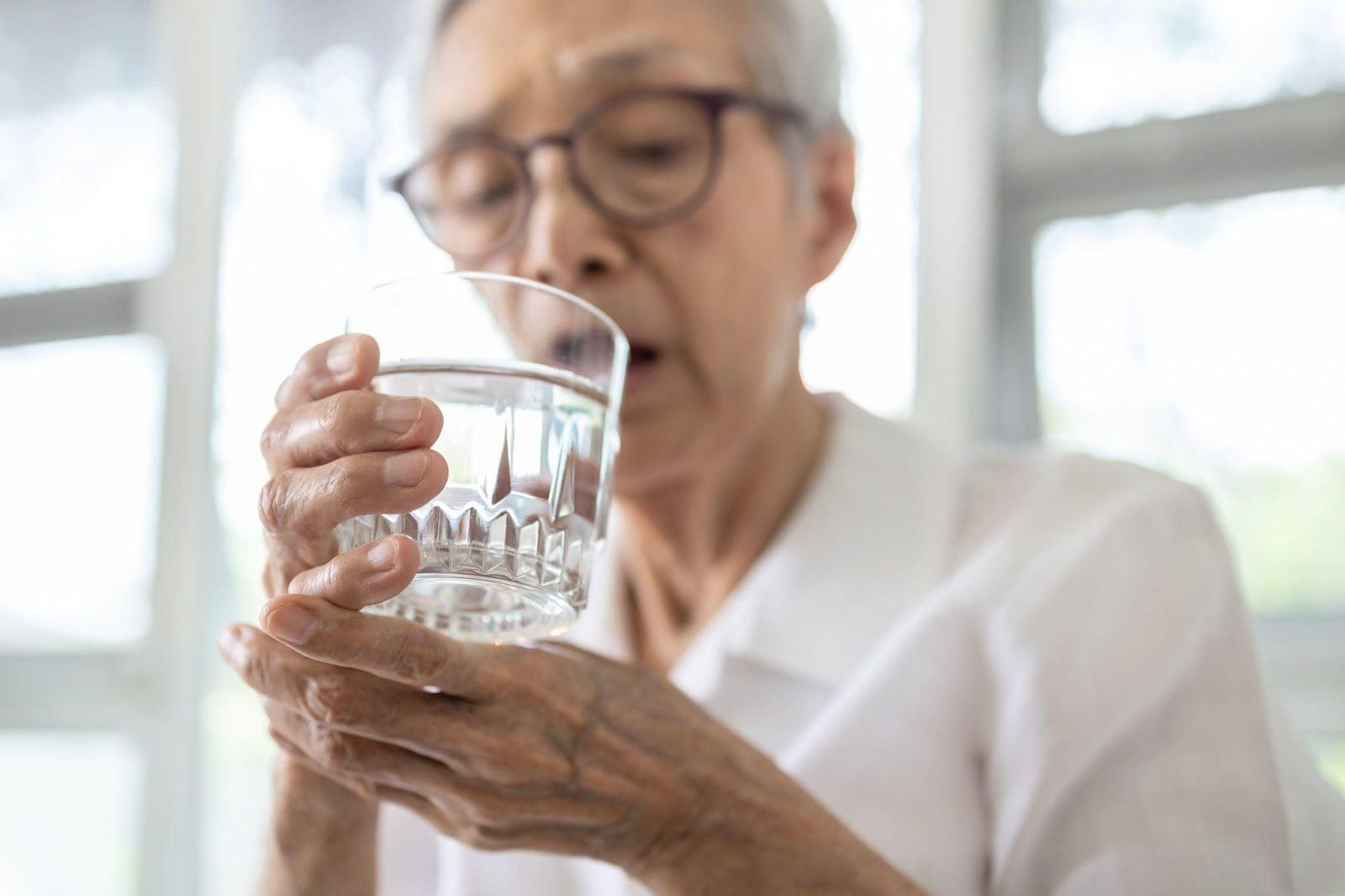 Senior woman steadying her shaky hand while drinking a glass of water.