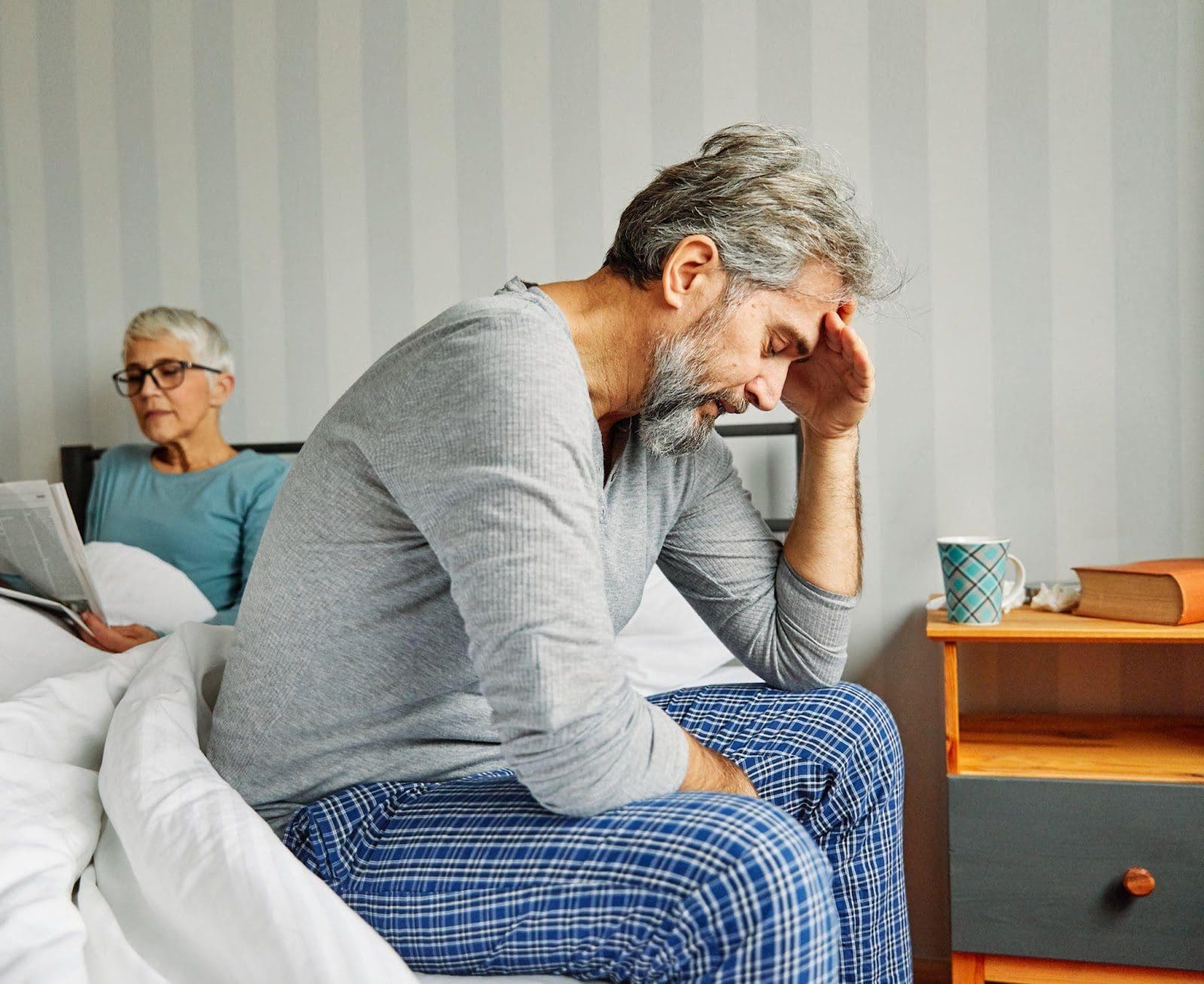 Unhappy senior man struggling with erectile dysfunction sitting on the edge of bed while his wife reads a book.