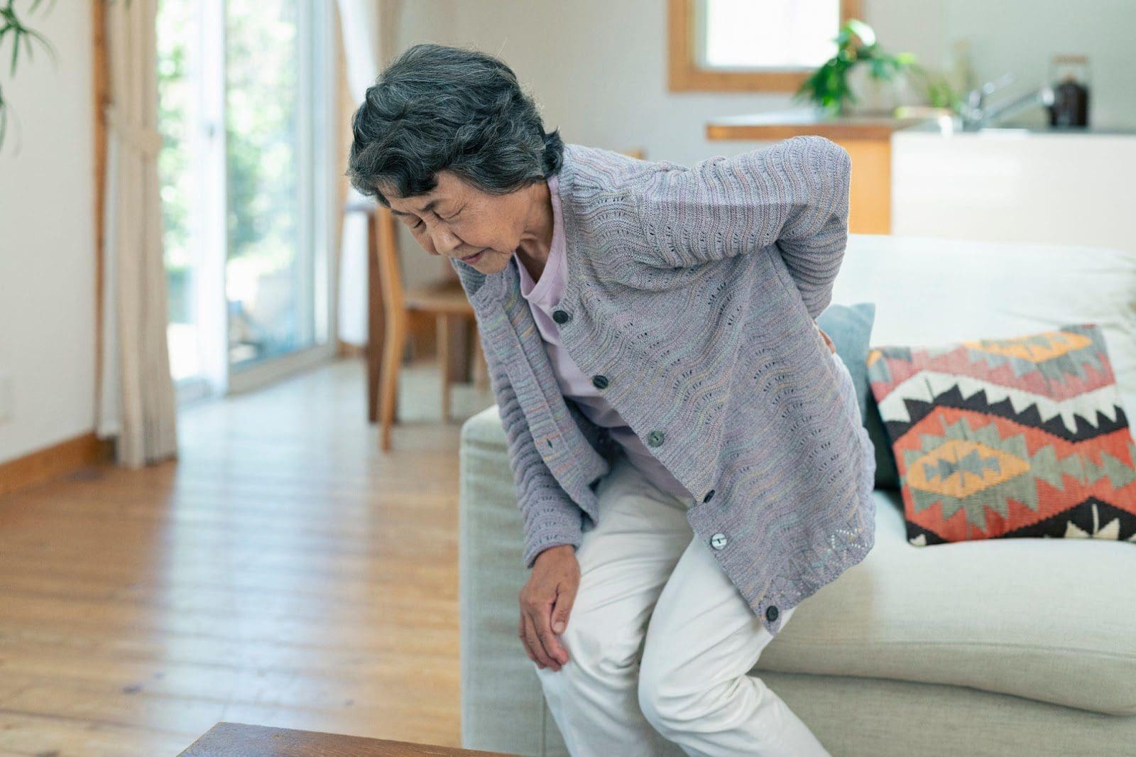 Senior woman standing next to a couch, bending over and placing her hand on her lower back in pain.